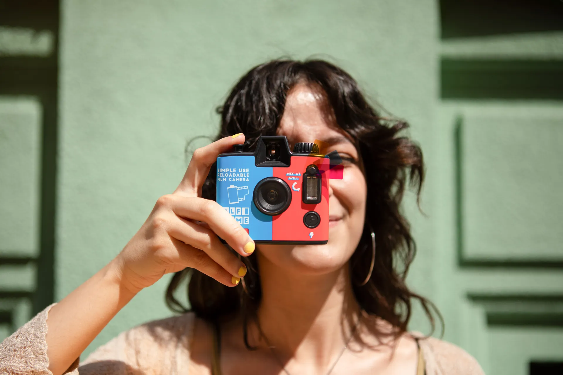 A colorful disposable camera held up to a face, ready to take a photo, with a green wall in the background.